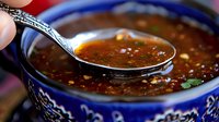 Spicy red chili sauce with garlic and herbs in decorative blue ceramic bowl, silver spoon scooping aromatic Asian dipping sauce with visible chili flakes and oil.
