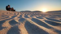 Rippled sand dunes at sunset with silhouetted couple sitting on top, warm golden light casting long shadows across desert landscape creating natural patterns in sand.