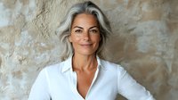 Confident mature woman with silver gray hair wearing white shirt smiling at camera against textured wall background. Natural aging and beauty concept for lifestyle marketing.