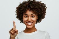 Happy young African American woman with curly afro hair pointing finger up showing number one gesture, smiling at camera in white casual shirt.