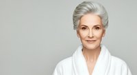 Mature woman with silver hair wearing white robe against neutral background, suitable for skincare, wellness, beauty and anti-aging product marketing campaigns.