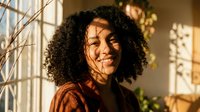 Young African American woman with curly hair smiling in warm sunlight streaming through window, creating natural shadows and cozy indoor atmosphere.