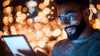 Young African American man in glasses smiling while using digital tablet at night, illuminated by warm bokeh lights creating magical atmosphere. Cozy evening technology use.