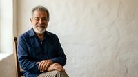Mature man with gray beard and salt-and-pepper hair, appearing to be of Mediterranean or Middle Eastern descent, wearing a navy blue button-up shirt, seated indoors against a white textured wall with natural window lighting.