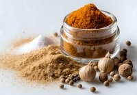 Aromatic spices collection with turmeric powder in glass jar, garlic, nutmeg, and peppercorns on white background for cooking and seasoning.