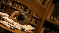 Young male student sleeping on books at library desk surrounded by bookshelves during late night study session for academic research and exam preparation.