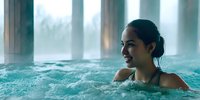 Young Asian woman relaxing in indoor spa jacuzzi with steam rising from water, smiling while enjoying hydrotherapy treatment at luxury wellness resort.