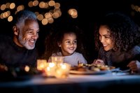 African American family enjoying candlelit dinner together, senior man with granddaughter and mother sharing warm moments with bokeh lights in background.