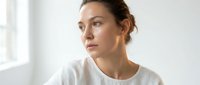 Young woman with brown hair in casual white t-shirt looking thoughtfully to the side near bright window, suitable for lifestyle, wellness, and portrait projects.