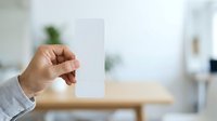 Hand holding blank white rectangular card in modern office environment with wooden desk and blurred background, suitable for branding, mockups, and business presentations.