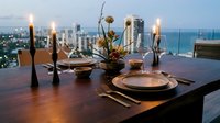 Elegant rooftop dining setup with lit candles, floral centerpiece, and place settings overlooking modern cityscape and ocean at sunset for upscale event planning.