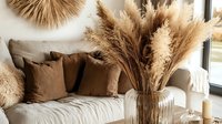 Cozy boho living room interior with pampas grass in vase, beige sofa, brown cushions, and natural decor elements creating warm minimalist atmosphere.