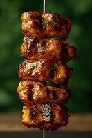 Grilled chicken kebab with glazed marinade and herbs on metal skewer against dark green background, close-up shot showing juicy, caramelized meat texture.