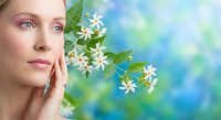 Young Caucasian woman with natural makeup touching face near white jasmine flowers on soft blue green bokeh background for skincare and beauty concepts.