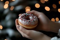 Hands holding festive glazed donut with red and green sprinkles against warm bokeh lights background for holiday celebrations and seasonal treats.