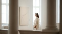Young woman in cream sweater contemplating minimalist line art in bright modern gallery space with natural window light and neutral tones.