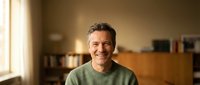 Middle-aged Caucasian man smiling warmly in cozy home interior wearing green sweater. Natural lighting creates inviting atmosphere for lifestyle content.