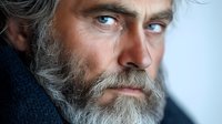 Close up portrait of mature Caucasian man with gray beard and striking blue eyes against light background, showing contemplative serious expression.
