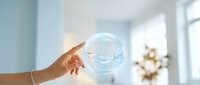 Young woman holding transparent soap bubble with water droplets against bright window and blurred interior background, symbolizing fragility, purity, and environmental awareness.