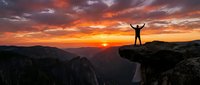 Hiker celebrating success on mountain peak at dramatic sunset with arms raised, overlooking canyon landscape with vibrant orange and purple sky clouds.