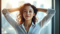Young Asian woman with curly hair and white shirt smiling with hands behind head near window in morning sunlight, expressing joy and relaxation in natural lighting.