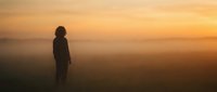 Silhouette of woman standing in misty field during golden hour sunrise, contemplating peaceful morning landscape with warm orange sky and fog.