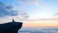 Silhouette of person sitting on mountain cliff edge above clouds during dramatic sunset sky with pink and blue colors. Contemplative solitary moment.