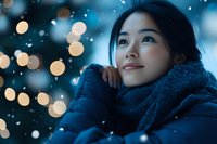 Young Asian woman in warm winter scarf gazing upward dreamily against blurred bokeh lights and falling snow, creating magical seasonal atmosphere.