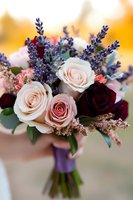 Romantic wedding bouquet featuring blush pink and burgundy roses with purple lavender sprigs and delicate greenery, wrapped in violet ribbon against soft bokeh background.