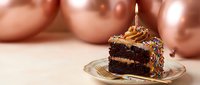 Chocolate birthday cake slice with lit candle and colorful sprinkles on glass plate against rose gold balloon backdrop for celebration party dessert.
