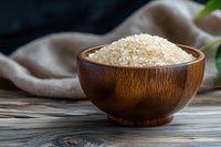 Raw white rice grains in wooden bowl on rustic table with natural linen napkin and green plant leaf, dark moody background for food photography.