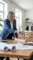 Mature female architect in blue blazer presenting wooden house model to client in bright office with blueprints and plants, real estate development consultation.