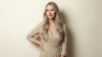 Young blonde Caucasian woman in beige wrap dress posing confidently against neutral background for fashion and lifestyle content.