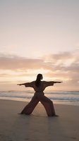 Woman practicing martial arts on beach at sunset with arms extended in warrior pose against golden sky and ocean waves.