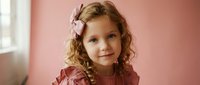 Young girl with curly blonde hair and pink bow wearing coral dress poses against soft pink background in natural window light for portrait photography.