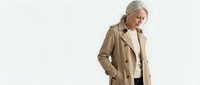 Senior woman with gray hair wearing a beige trench coat and cream blouse, standing against white background with hand in pocket, suitable for business and lifestyle marketing.