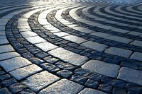 Curved cobblestone pavement pattern with sunlight creating dramatic shadows and highlights on stone blocks, forming concentric arcs in urban setting.