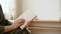 Young adult male with light skin tone reading an open book indoors near a window with natural light, suitable for education, learning, and lifestyle content.