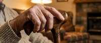 Senior man with wrinkled hands holding wooden cane indoors, representing aging, mobility assistance, healthcare, elderly care, and retirement lifestyle concepts.