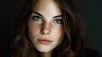 Close-up portrait of young woman with freckles and green eyes against dark background, showcasing natural beauty and authentic expression.