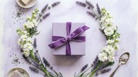 Purple gift box with ribbon surrounded by lavender flowers and white blooms on soft background. Elegant present arrangement for special occasions and celebrations.