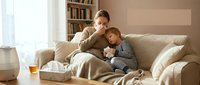 Father and young son sitting together on beige sofa, sharing warm moment while reading book. Cozy home interior with bookshelf and natural light. Family bonding and parenting concept.