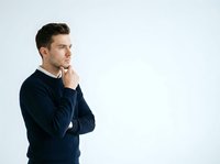 Thoughtful young man with dark hair and light skin wearing navy sweater, hand on chin in contemplative pose against white background for business and lifestyle concepts.