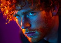 Young male model with ginger hair in dramatic orange and blue neon lighting against dark background, close-up portrait showing intense gaze and facial features.