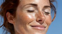 Young woman with freckles and closed eyes smiling against blue sky background, enjoying sunshine on her face.