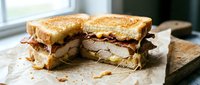Grilled turkey and bacon sandwich with melted cheese on toasted bread, cut in half showing layers, served on parchment paper for lunch or dinner meal.