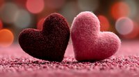 Two textured fabric hearts in burgundy and pink sitting together on glittery surface with warm bokeh lights creating romantic Valentine's Day atmosphere.