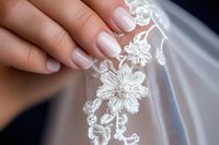 Delicate female hands with french manicure touching white floral lace fabric, close-up view showing intricate flower pattern details and soft tulle texture. Perfect for wedding themes.