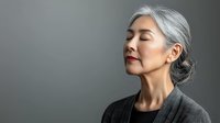 Serene mature Asian woman with gray hair in bun wearing gray sweater, eyes closed with peaceful expression against neutral background. Portrait conveys wisdom and mindfulness.