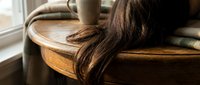 Young woman with long brown hair relaxing on wooden chair by window in cozy home interior with warm natural lighting and soft textiles.